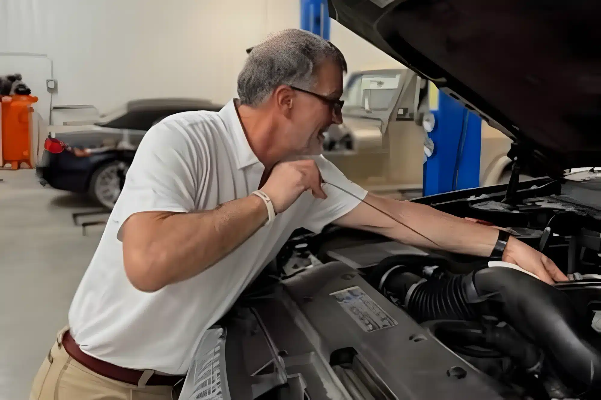 A man in a white polo shirt checks the oil level of a car engine in a garage, holding the dipstick and looking closely while the hood is open. Other vehicles and equipment are visible in the background.