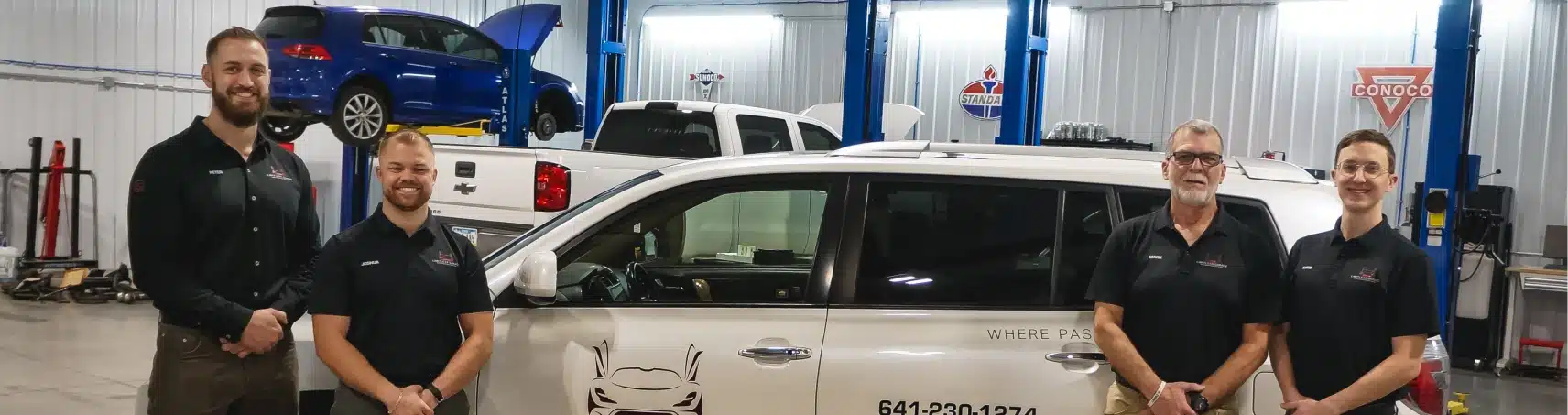 Four men in black shirts stand smiling next to a white vehicle inside an auto repair shop, with a blue car and a white truck elevated on lifts in the background.