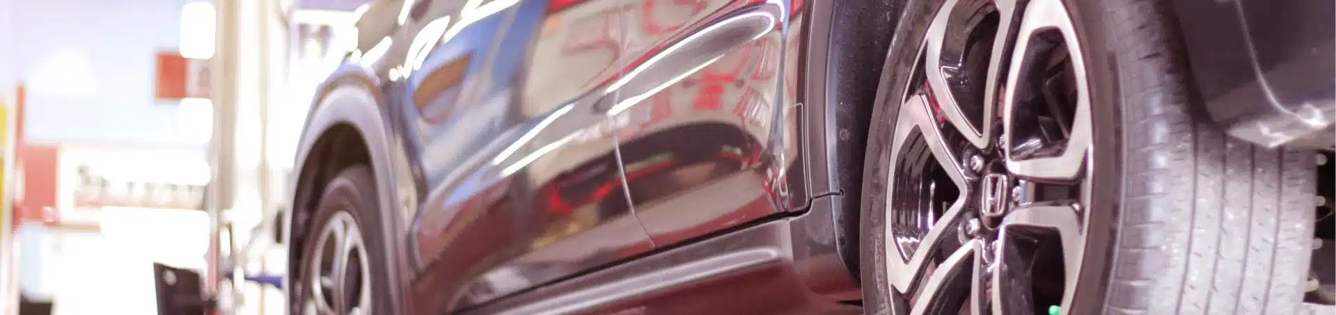 A close-up side view of a black car parked indoors, focusing on the rear tire and wheel with the car’s glossy exterior reflecting light. The background is blurred, showing some workshop equipment.