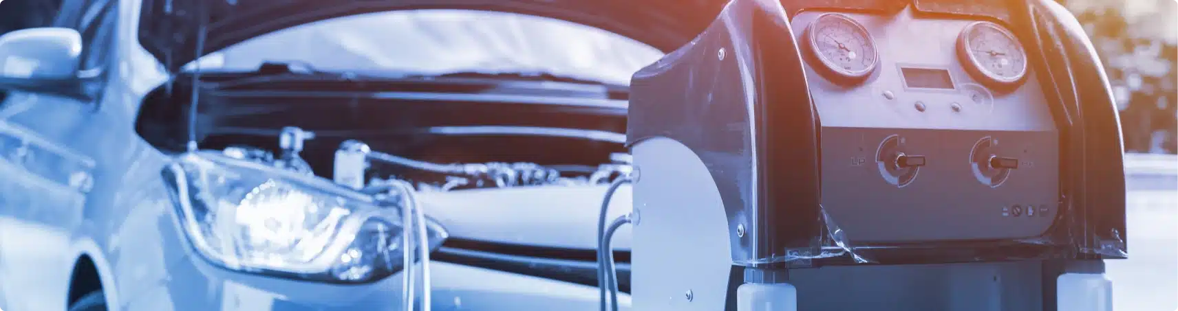 A car with its hood open is parked in the background, while an automotive air conditioning service machine with gauges is in focus in the foreground.