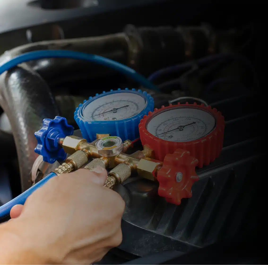 A persons hand adjusts a manifold gauge set with blue and red dials, used for measuring pressure in an automotive air conditioning system.