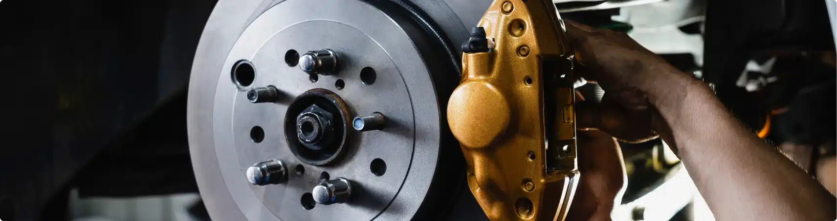 A close-up of a person’s hands working on a car’s brake system, showing a shiny metallic brake disc and a gold-colored brake caliper being adjusted or installed.