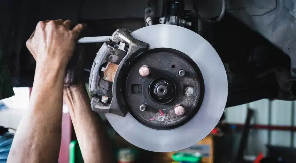 A person’s hands are working on a car’s exposed disc brake system, using a tool to adjust or repair the brake caliper. The surroundings suggest a garage or auto repair shop setting.