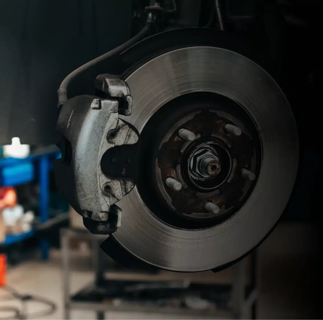 Close-up view of a car brake disc and caliper mounted on a wheel hub, with a workshop bench and tools blurred in the background.
