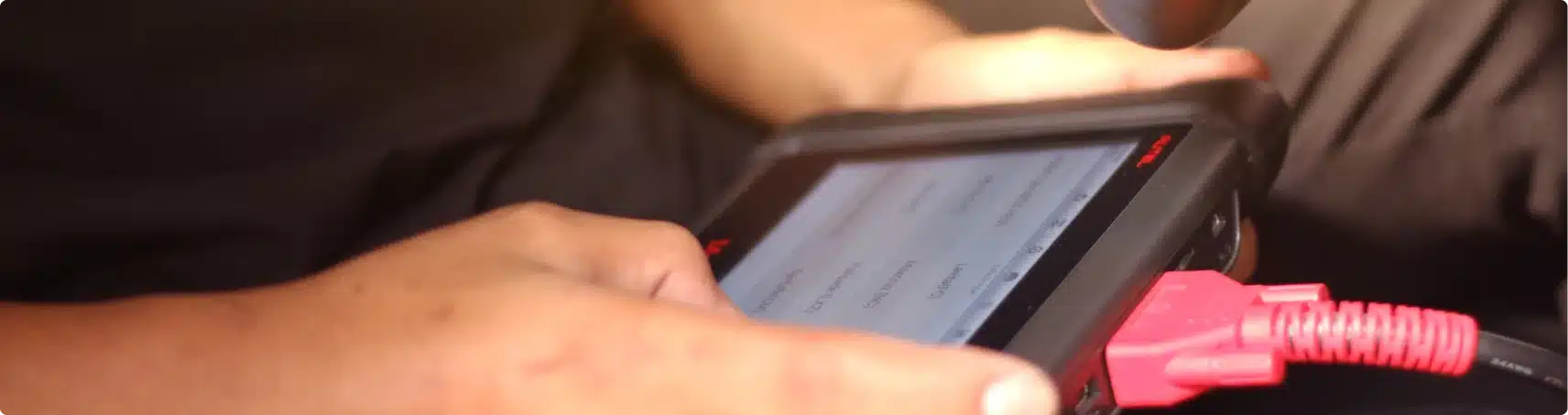 Close-up of a person’s hands using a diagnostic tablet device with a red cable attached, likely for vehicle diagnostics. A screen displays menu options, and the background is out of focus.