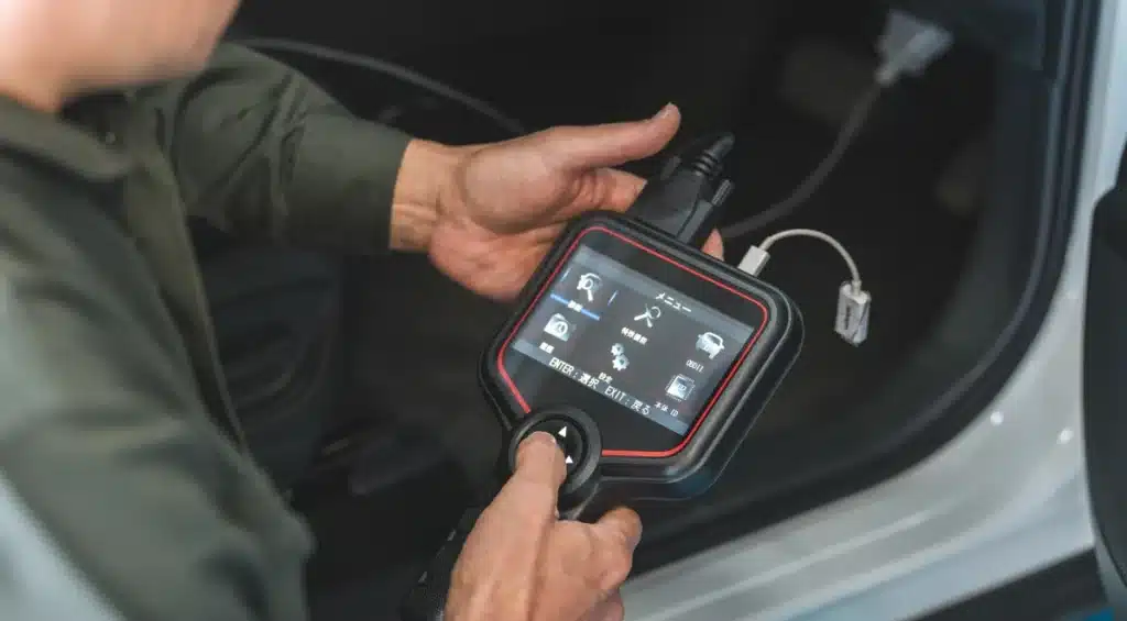 A person holds and operates a car diagnostic tool with a touchscreen interface, connected to a vehicles onboard port inside a car.