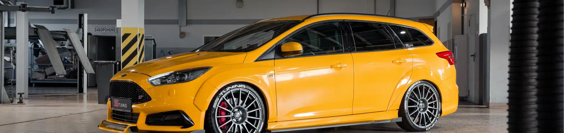 A bright yellow Ford Focus station wagon with sporty black rims is parked inside a modern, well-lit garage or workshop. The car has red brake calipers and tinted windows.