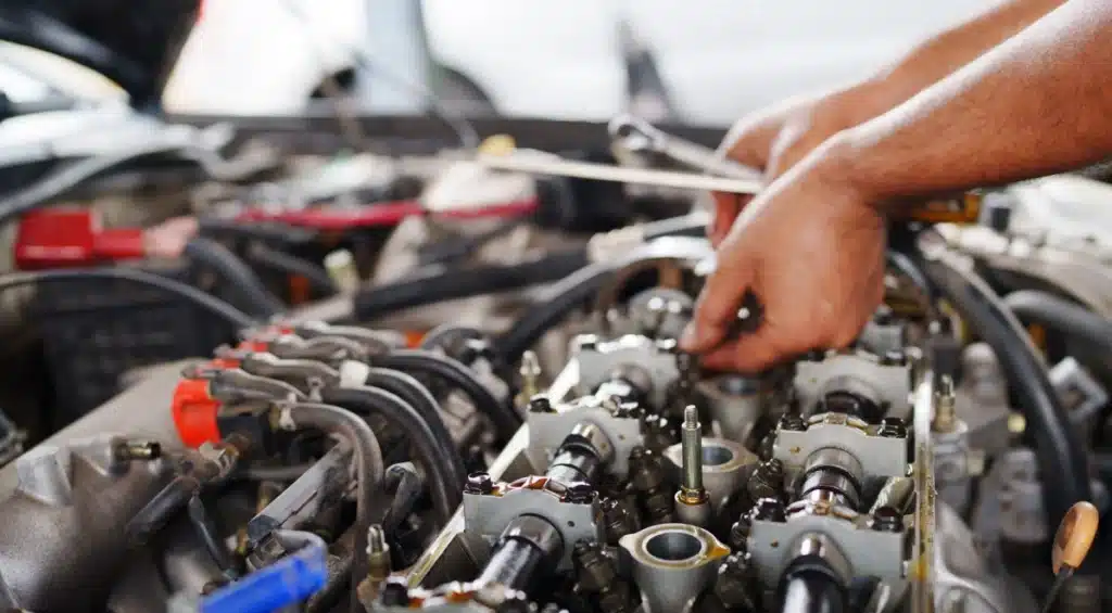 Close-up of a person’s hands working on a car engine, using tools to adjust components under the hood, with various engine parts and wiring visible.