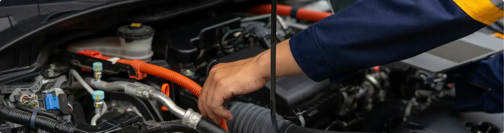 A person wearing a blue and yellow uniform works on a car engine, reaching inside the open hood near orange cables and various engine components.