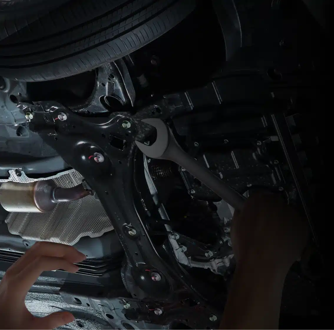A person is using a wrench to work on the underside of a car, likely performing maintenance or repairs near the exhaust system.