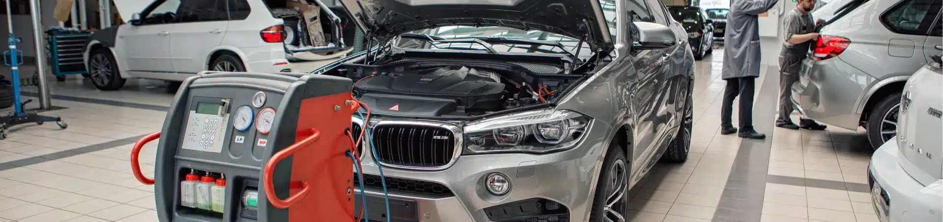 A silver BMW SUV with its hood open is connected to diagnostic equipment in an auto repair shop. Other cars and two mechanics in lab coats working in the background are visible.
