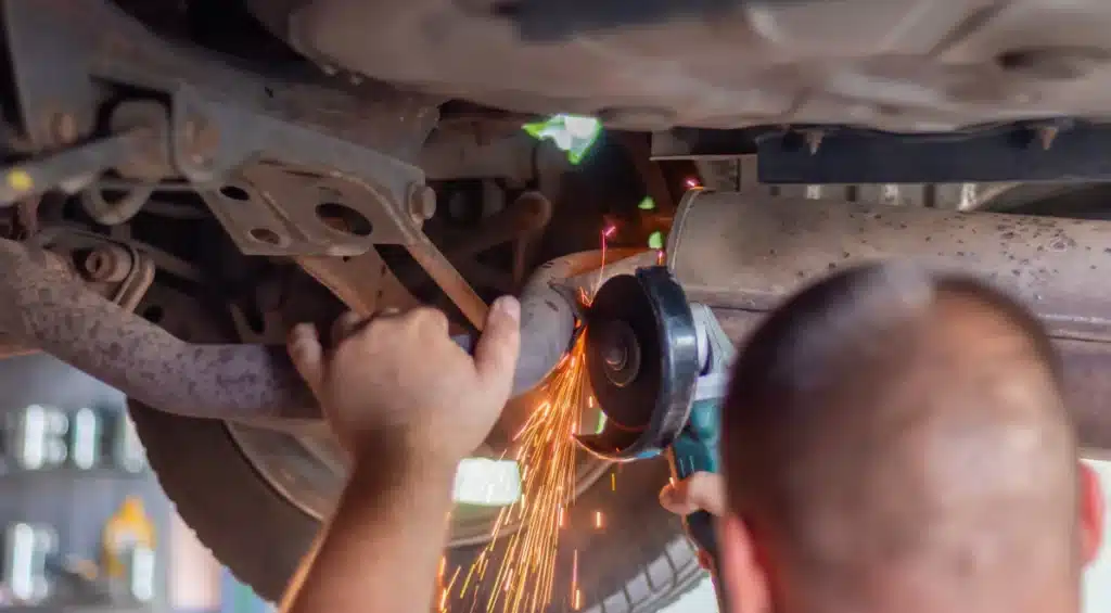 A person uses an angle grinder to cut a metal exhaust pipe under a vehicle, creating bright sparks. The underside of the vehicle and various car parts are visible.