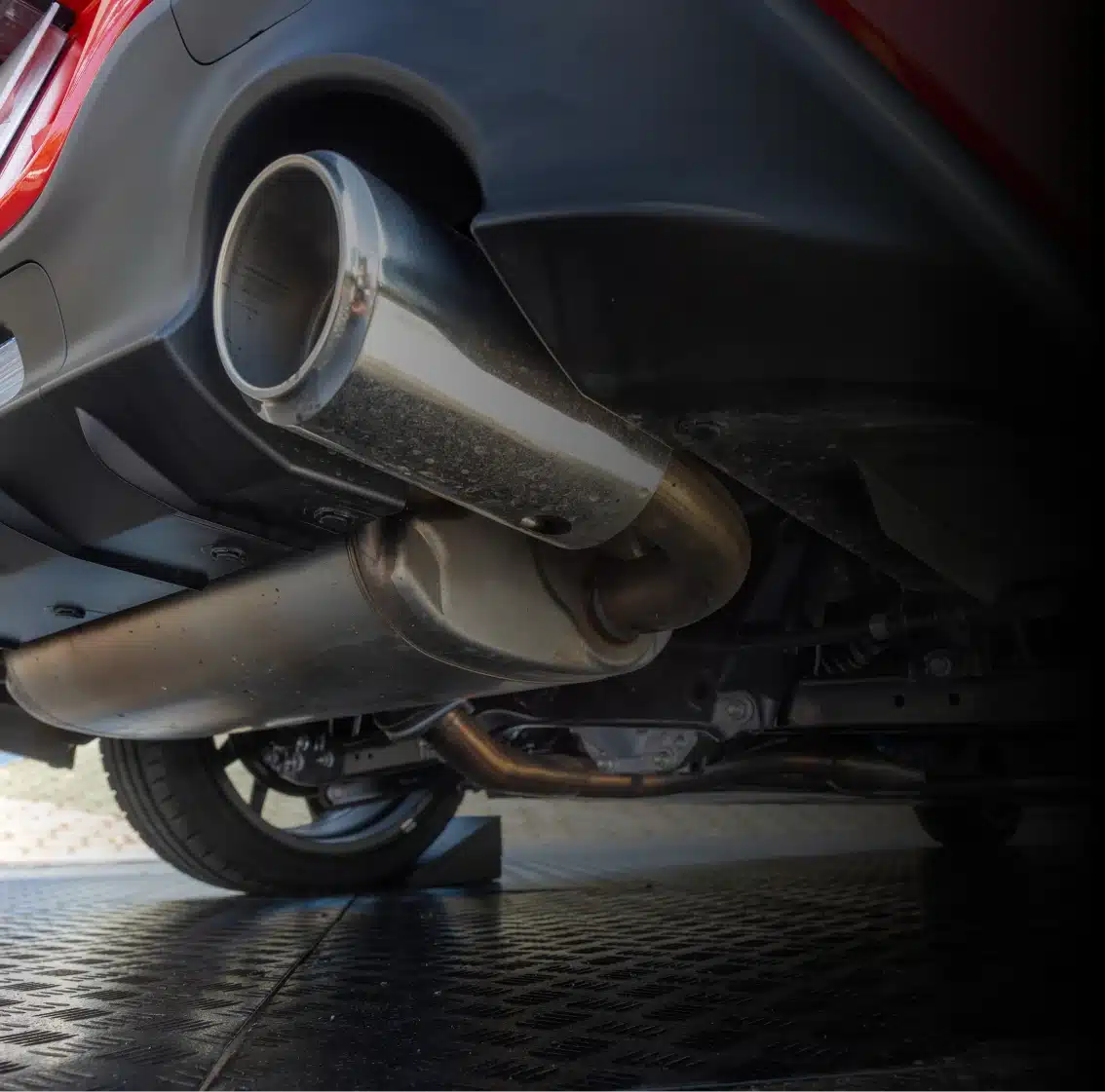Close-up of a car’s dual exhaust pipes underneath the rear bumper, showing metallic details and part of the rear wheel, captured on a reflective surface.