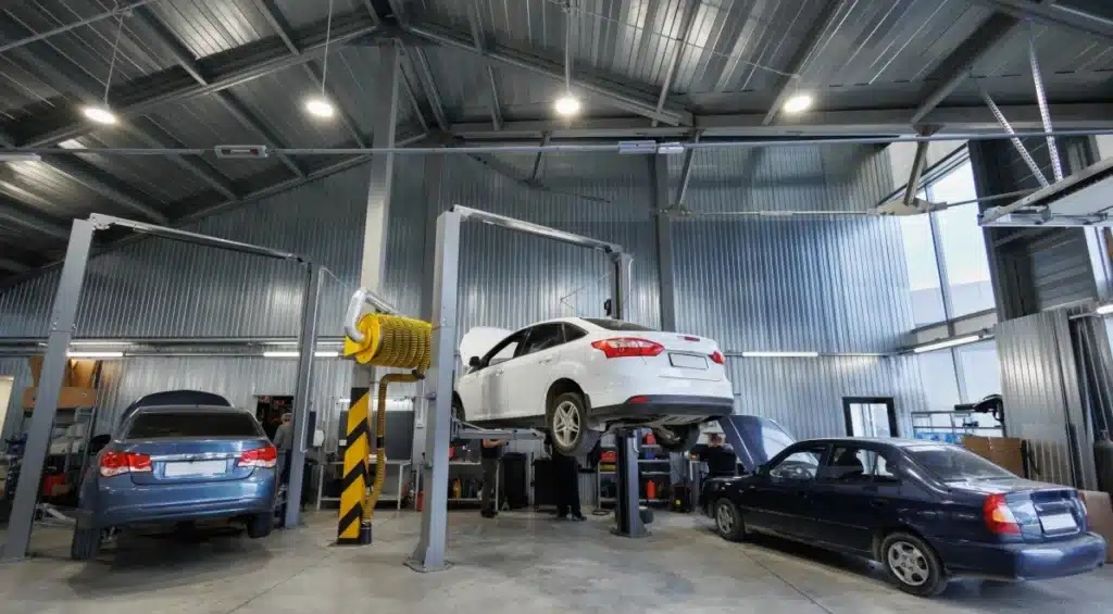 Several cars in a modern auto repair shop, with one white car raised on a hydraulic lift and mechanics equipment visible around the spacious, well-lit garage.