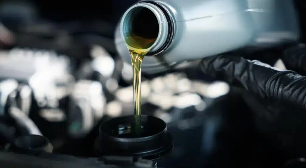 Close-up of a gloved hand pouring motor oil from a plastic bottle into a car engine, with the golden liquid flowing smoothly. The engine components are blurred in the background.