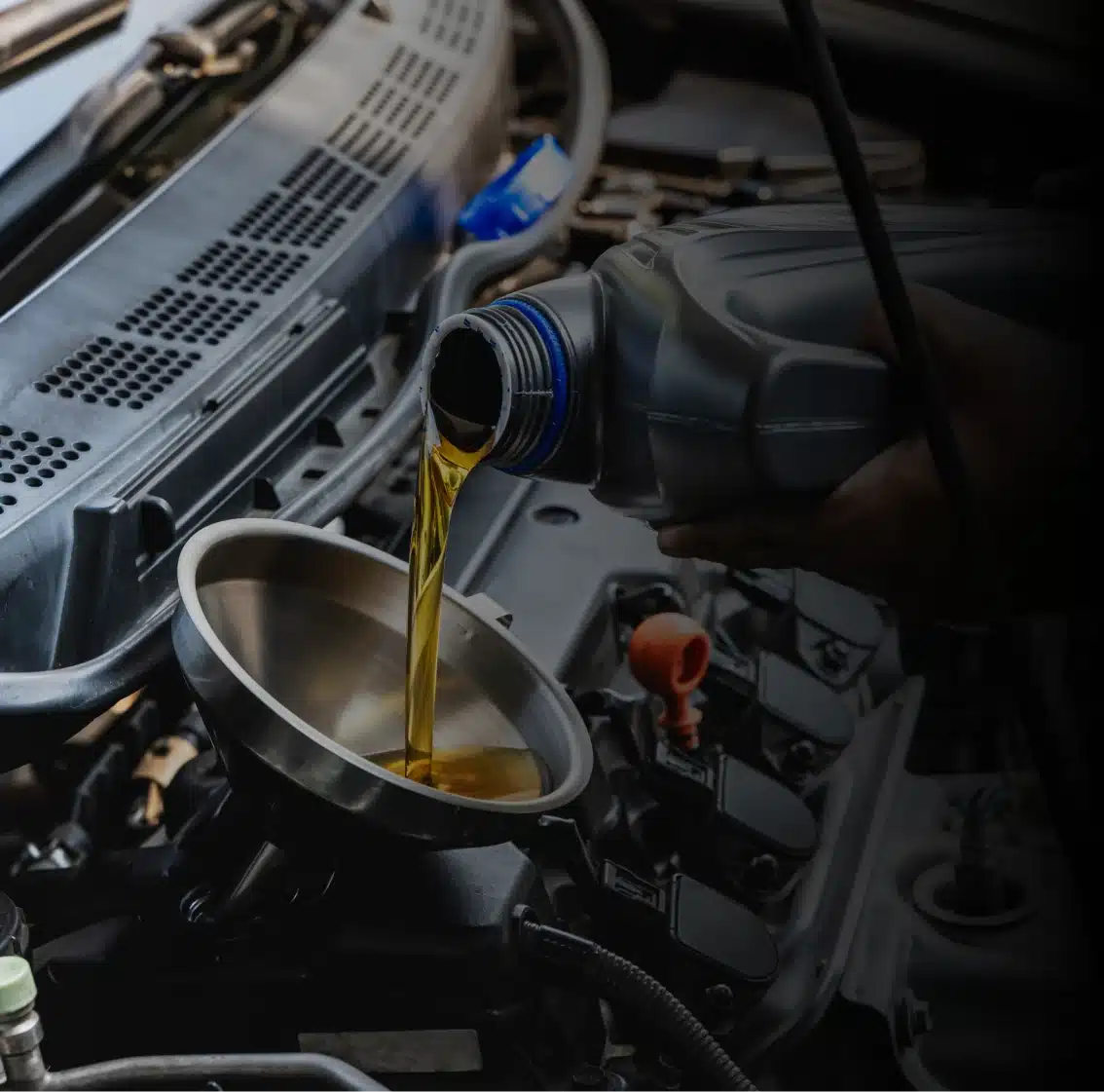 A person pours engine oil from a plastic bottle into a funnel placed in a car engine bay. The metal funnel ensures the oil flows directly into the engine without spilling.