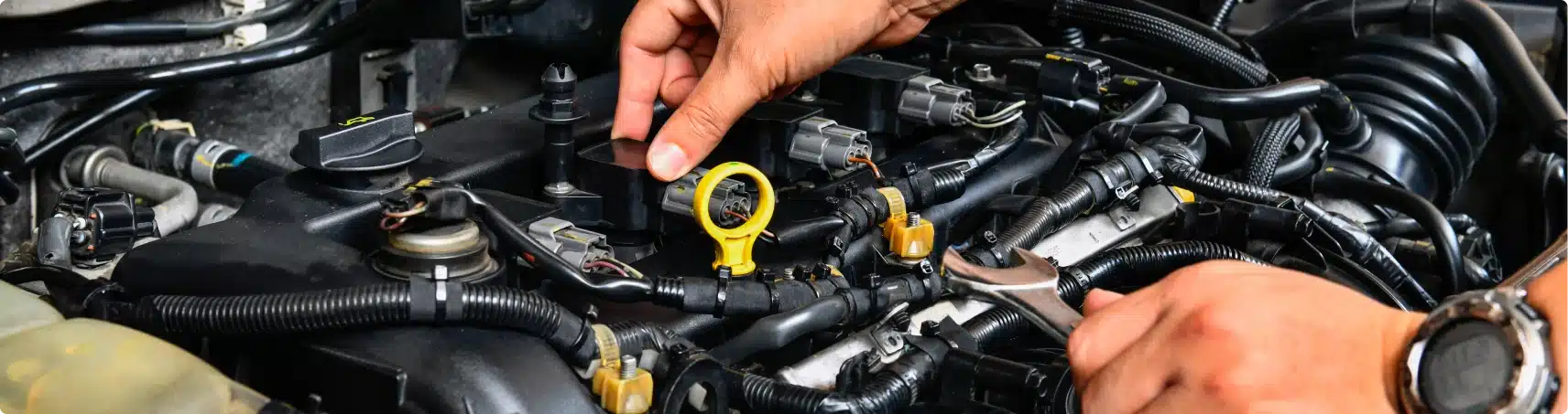 A close-up of hands working on a car engine, using a wrench to adjust components. Various wires, connectors, and engine parts are visible.