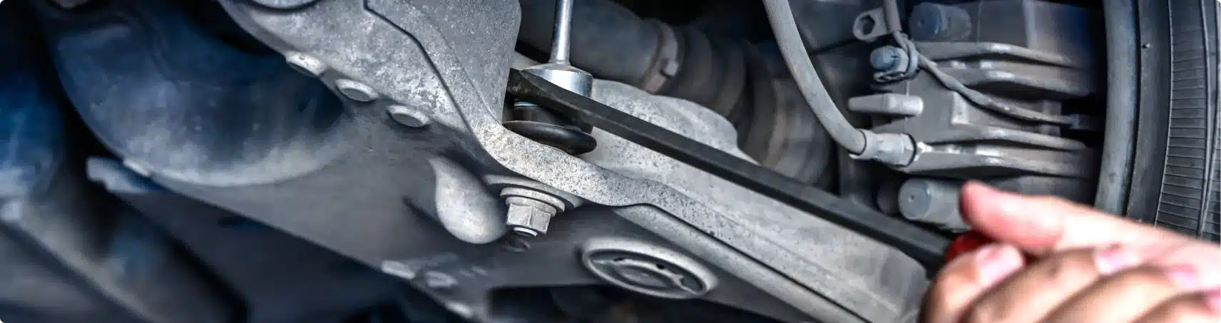 A close-up of a persons hand using a wrench to tighten a bolt on a cars suspension or steering system, showing metal parts and the wheel in the background.