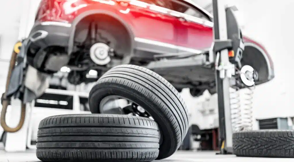 A red car is lifted on a service platform in an auto repair shop. Two tires are positioned in the foreground, and the car’s wheels have been removed for maintenance.