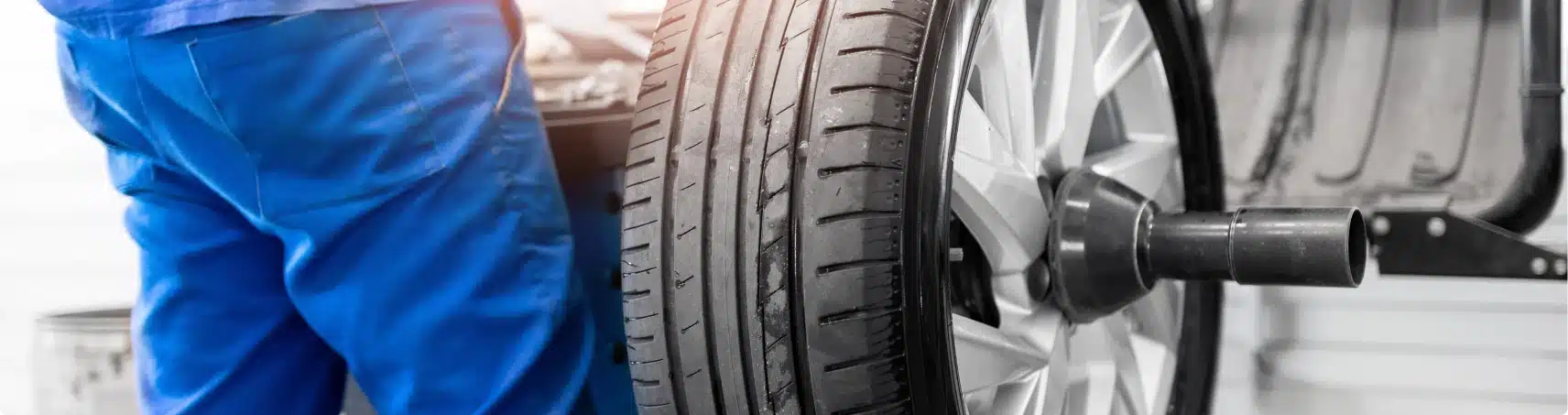 A mechanic in blue overalls stands next to a car tire mounted on a balancing machine in a workshop. The focus is on the tire and wheel, with the mechanic partially visible.