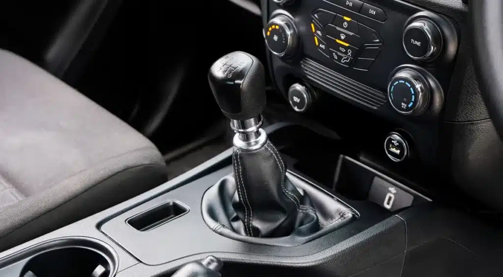 Close-up of a cars manual gear shift lever with black leather cover, surrounded by cup holders and dashboard controls for air conditioning and entertainment.