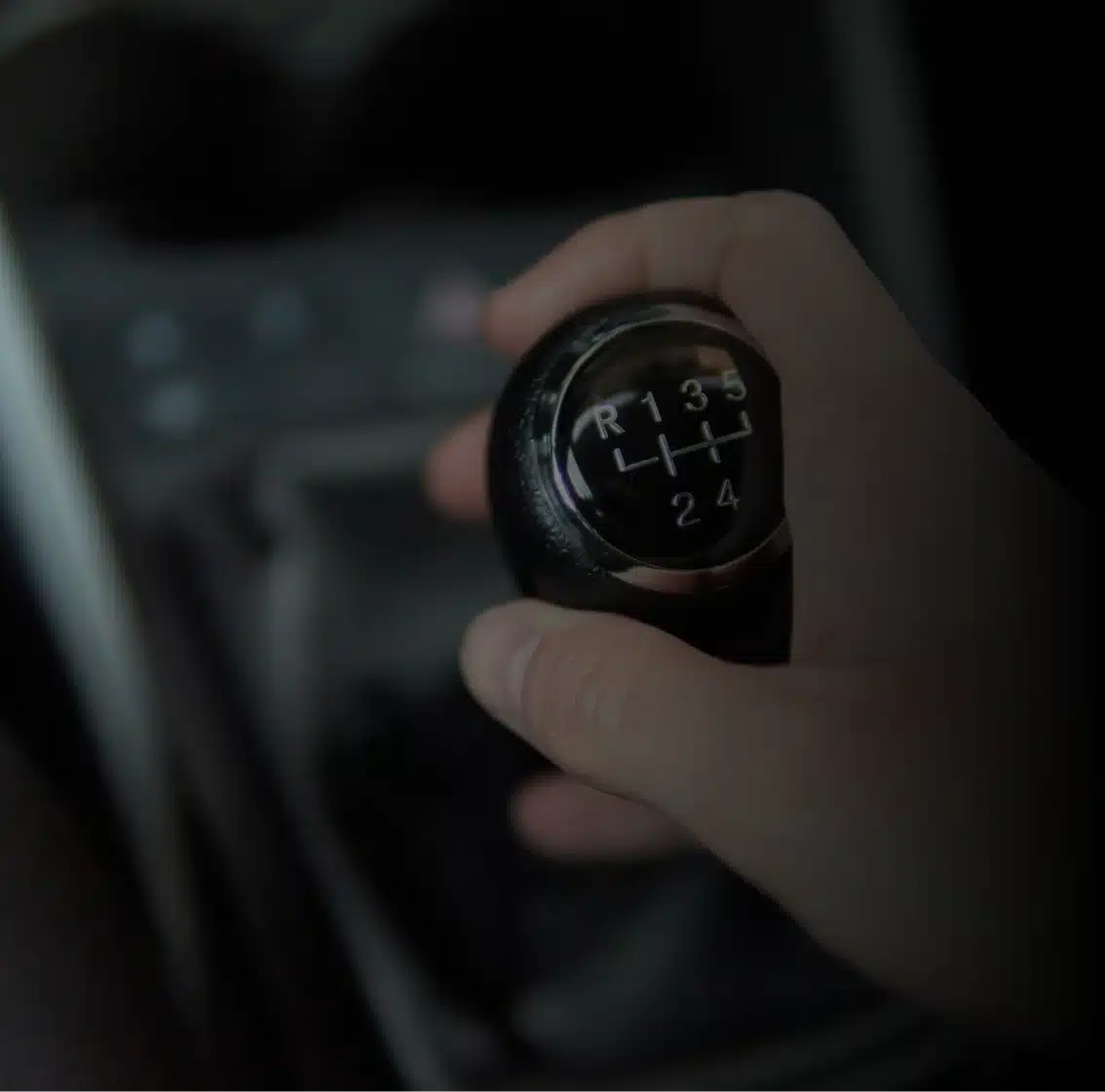 A hand gripping the gear shift of a manual transmission car, showing the gear pattern labeled R, 1, 2, 3, 4, and 5. The background is softly blurred.