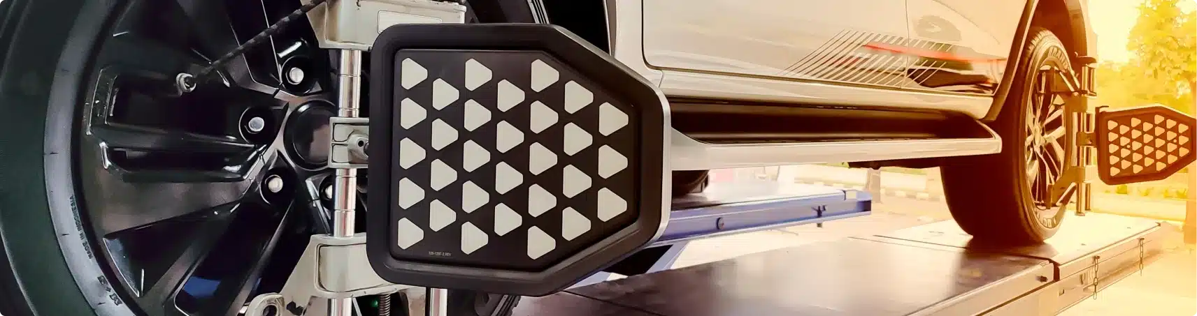 Close-up of a car undergoing wheel alignment, showing the alignment sensor attached to the front wheel and the side of the vehicle lifted on a platform in a brightly lit workshop.