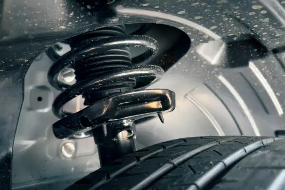 Close-up view of a car’s suspension system, showing a coiled spring and shock absorber above the tire inside the wheel well. The parts are metallic and slightly dusty.