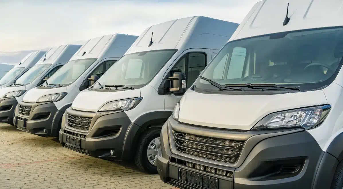 A row of white delivery vans is parked side by side on a paved lot, all facing the same direction under a bright sky, resembling a limitless garage ready for business.