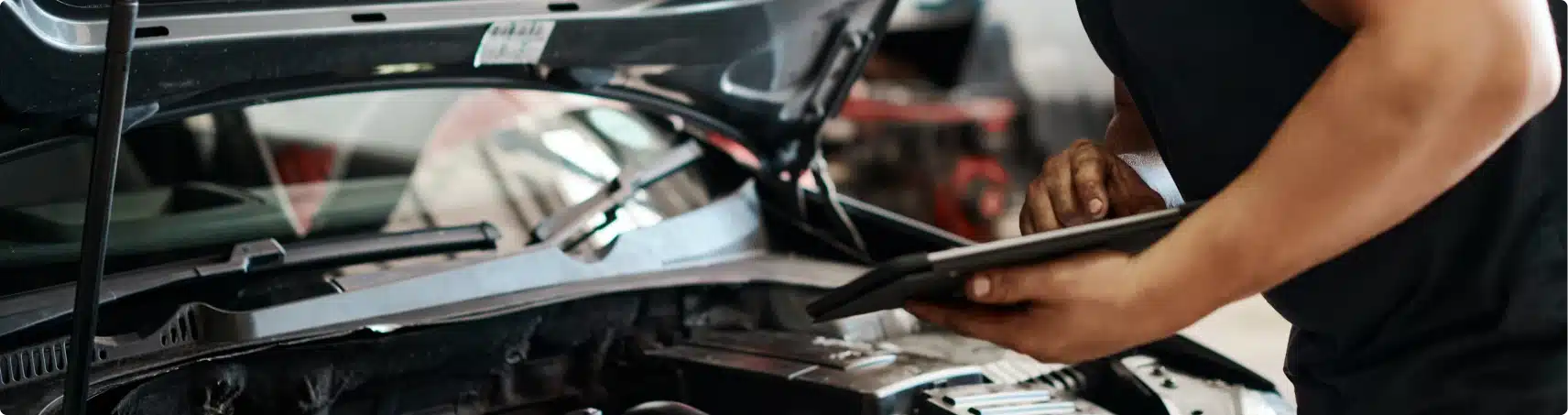 A person holds a tablet while inspecting a car engine with the hood open, appearing to perform diagnostics or Vehicle Inspections in an automotive workshop.